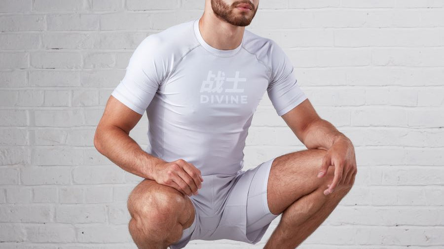 Man kneeling down wearing bjj rashguard and shorts. this man is in a stance as if he is watching his bjj instructor teach.