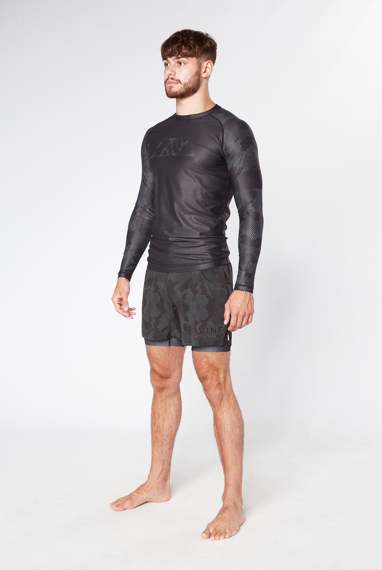A man stands barefoot, wearing a long-sleeved BJJ RASHGUARD by Divine JiuJitsu and matching shorts with subtle patterns. He has short hair and a beard and is gazing slightly to the side against a plain, light gray background.