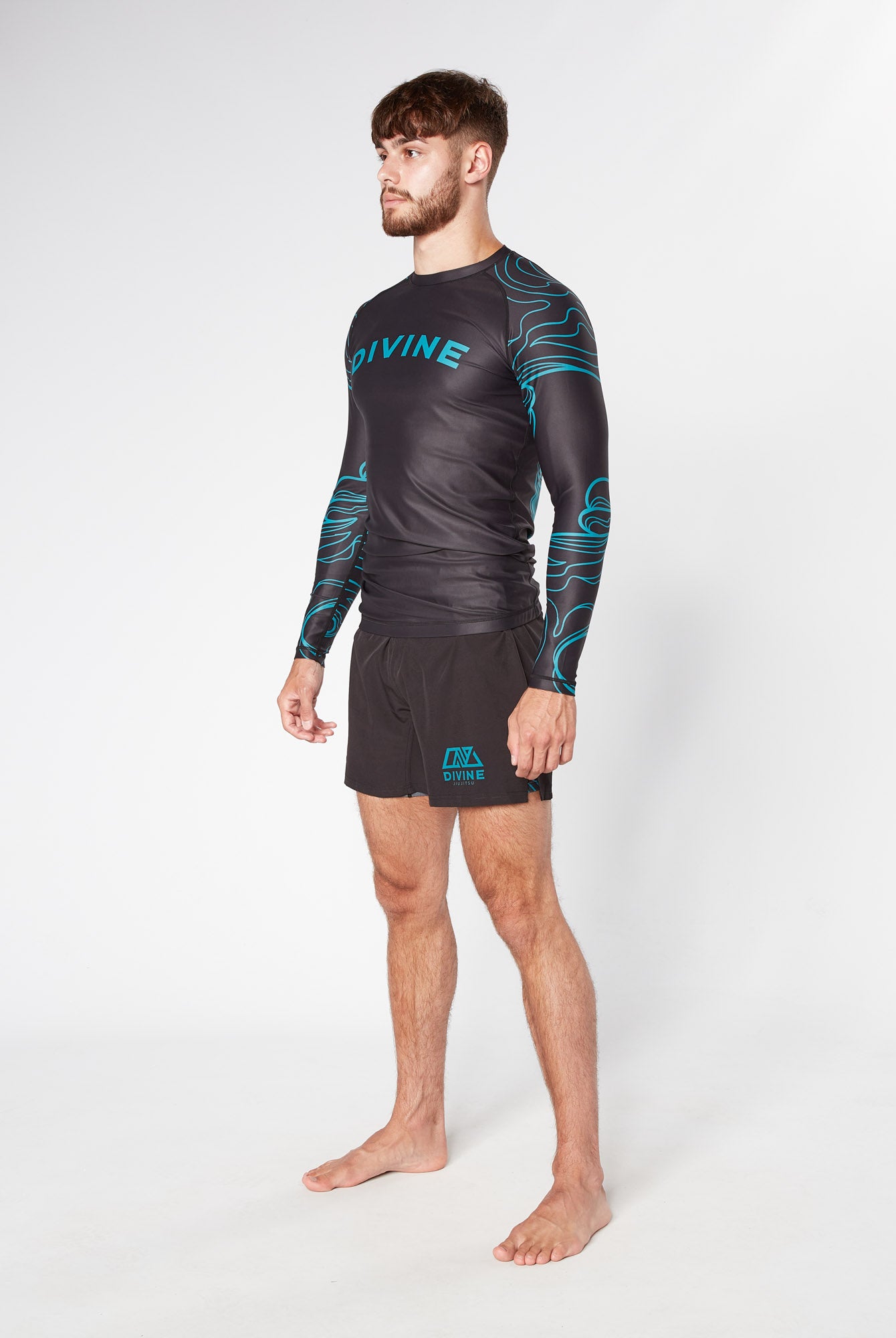 A barefoot individual stands against a plain white background. They are wearing the PHENOMENON RASHGUARD by Divine JiuJitsu, a long-sleeved black athletic shirt adorned with blue patterns and the brand name "DIVINE," made from moisture-wicking fabric. This is paired with black shorts that also feature a blue design. The person has short hair and a beard.