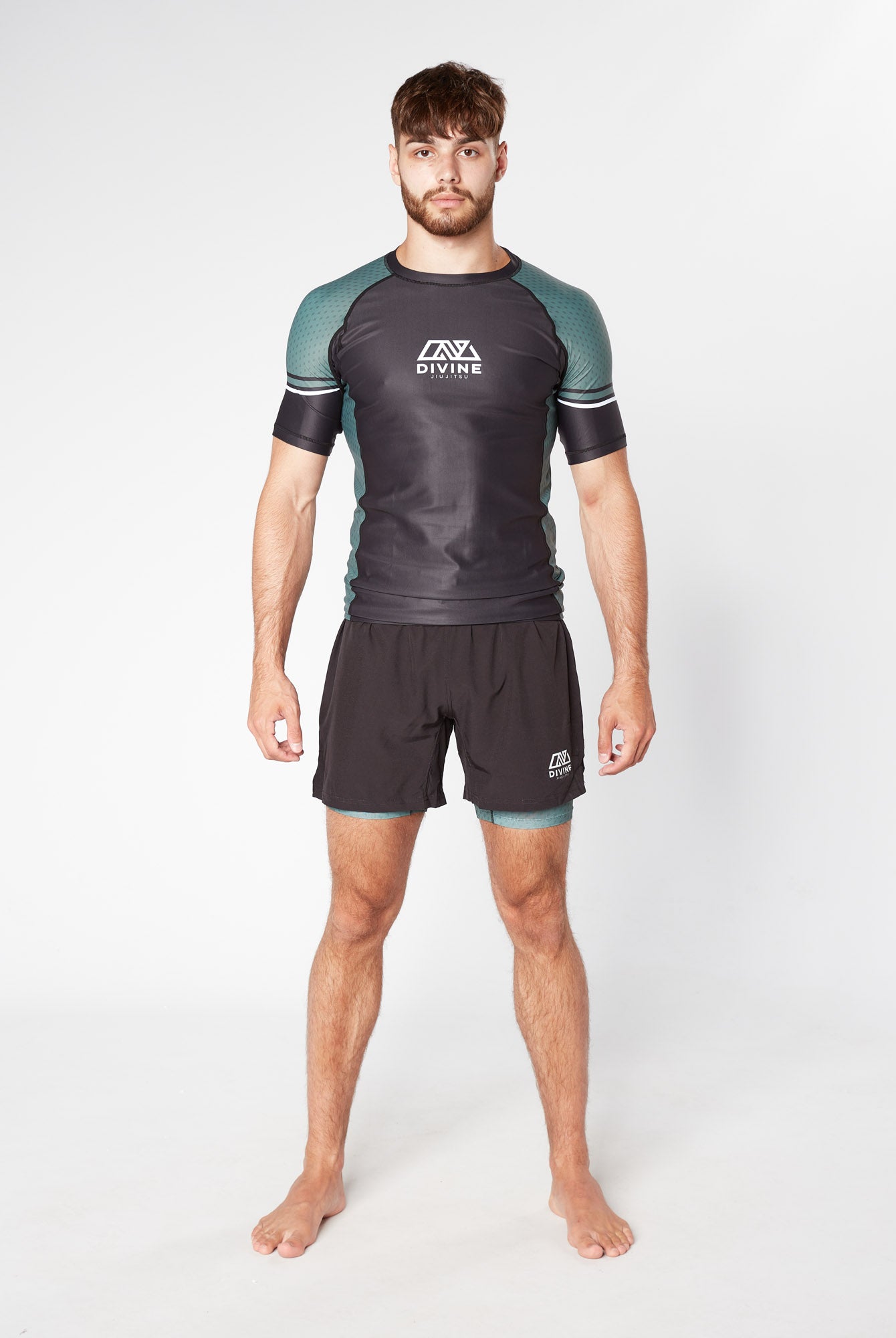A man stands barefoot against a plain white background, wearing a fitted athletic outfit featuring the LEGION KHAKI BJJ RASHGUARD from Divine JiuJitsu in black and green and matching black shorts. Made from moisture-wicking fabric, his attire ensures optimum performance. His arms are relaxed at his sides, and he has a neutral expression.