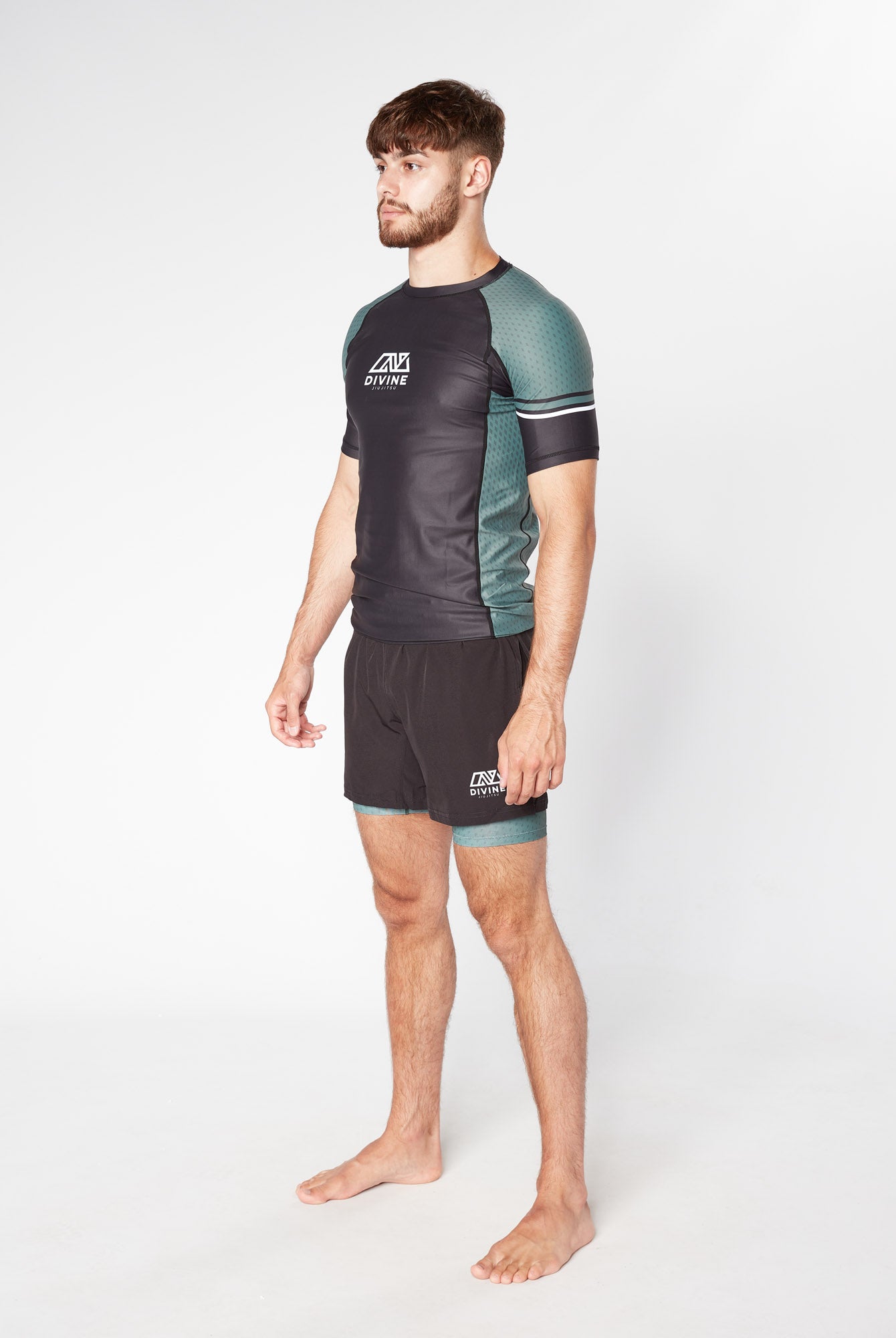 A bearded man with short hair stands barefoot in a studio against a plain white background, wearing the black and green LEGION KHAKI RASHGUARD by Divine JiuJitsu and matching shorts made of moisture-wicking fabric. He faces slightly to the side with a neutral expression.