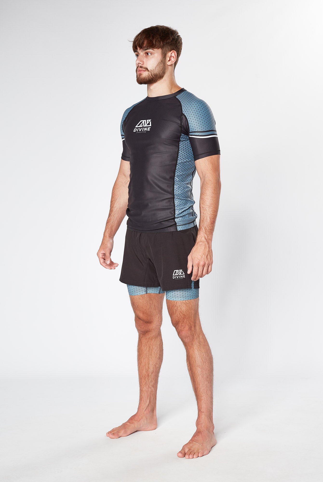 A barefoot man with a beard stands confidently, wearing a LEGION GREY RASHGUARD by Divine JiuJitsu, along with matching shorts. Both the shirt and shorts feature a hexagonal pattern and the logo "CV Fight Co." The background is plain white.