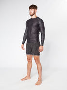 A man stands barefoot, wearing a long-sleeved BJJ RASHGUARD by Divine JiuJitsu and matching shorts with subtle patterns. He has short hair and a beard and is gazing slightly to the side against a plain, light gray background.