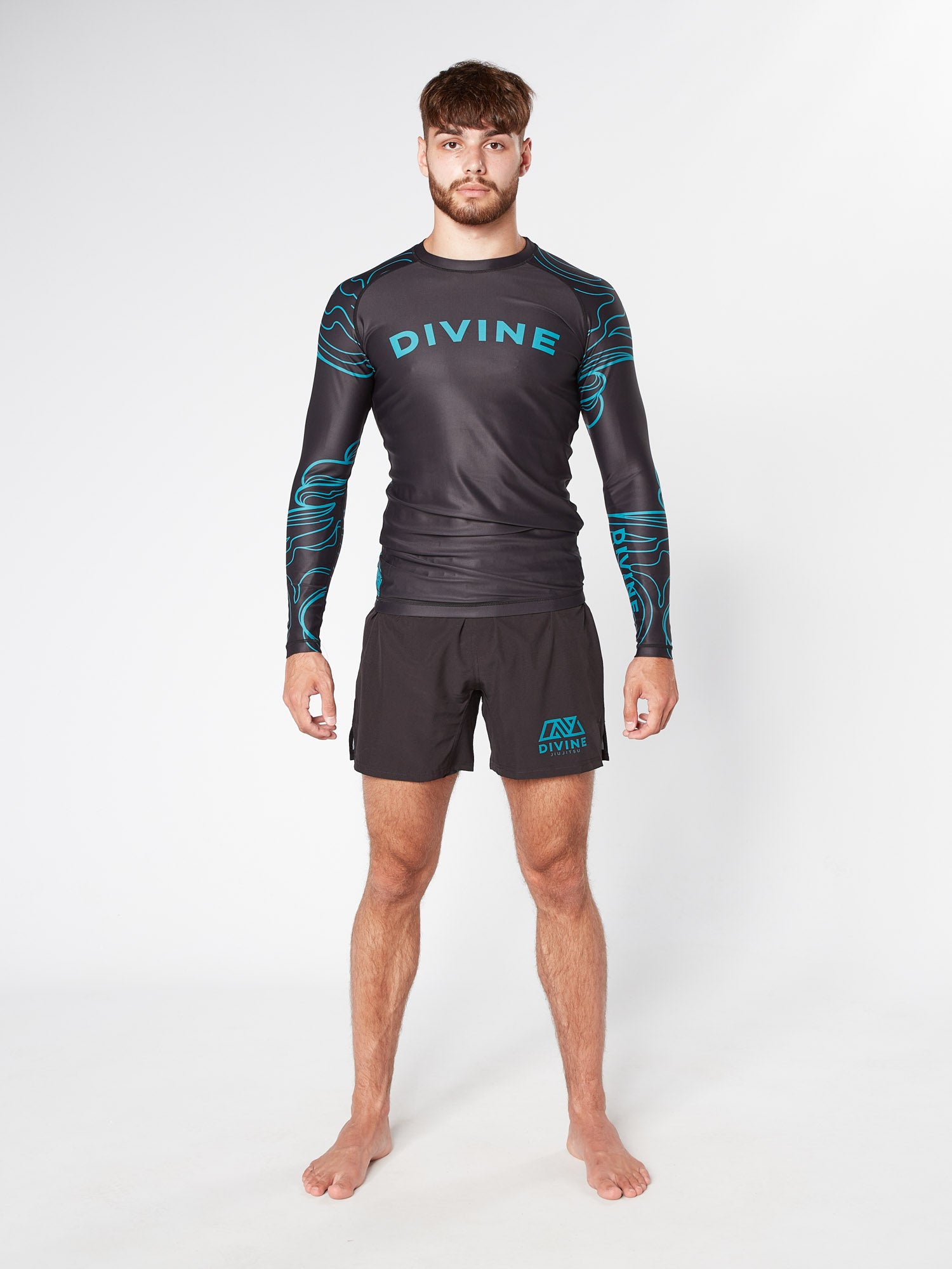 A barefoot man with short brown hair stands against a plain white background. He is wearing the PHENOMENON BJJ RASHGUARD, a black long-sleeved compression shirt with blue accents and the word "DIVINE" printed on the chest, resembling a sleek jiu-jitsu rashguard from Divine JiuJitsu. He pairs this with matching black shorts that feature a blue "DIVINE" logo on the left thigh.