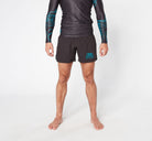 A person is standing barefoot against a plain white background, wearing a black long-sleeved rash guard with blue designs and Divine JiuJitsu's PHENOMENON BJJ  SHORTS featuring a small logo on the left thigh. The quick-dry fabric ensures comfort during intense sessions, but the model's face is not visible in the image.