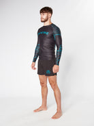 A barefoot individual stands against a plain white background. They are wearing the PHENOMENON RASHGUARD by Divine JiuJitsu, a long-sleeved black athletic shirt adorned with blue patterns and the brand name "DIVINE," made from moisture-wicking fabric. This is paired with black shorts that also feature a blue design. The person has short hair and a beard.