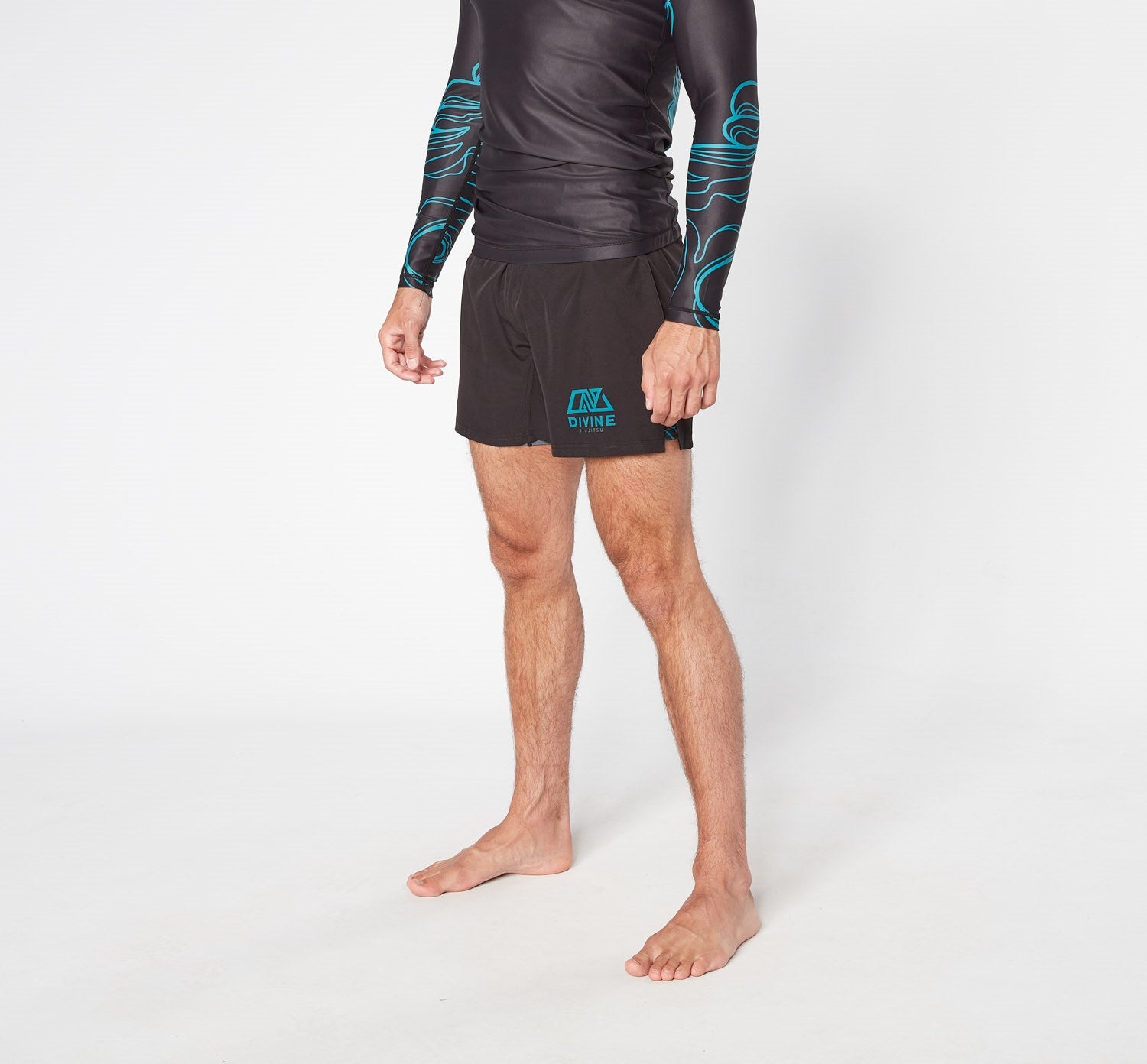 A person stands barefoot in a studio against a plain white background. They are wearing a black long-sleeve top with blue designs and the PHENOMENON SHORTS from Divine JiuJitsu, which feature black fabric with blue accents and quick-dry technology. Their head is not visible in the image.