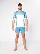 A man with short hair and a beard stands barefoot against a plain white background. He is wearing a light blue and white Divine JiuJitsu STEEL BJJ RASHGUARD and matching blue shorts. His arms are by his sides, and he looks directly at the camera.