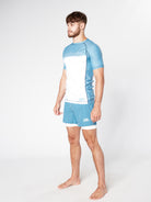 A man with short brown hair and a beard stands barefoot against a plain background, wearing the light blue and white STEEL RASHGUARD and matching shorts from Divine JiuJitsu. He is seen in a side profile, his gaze directed forward.