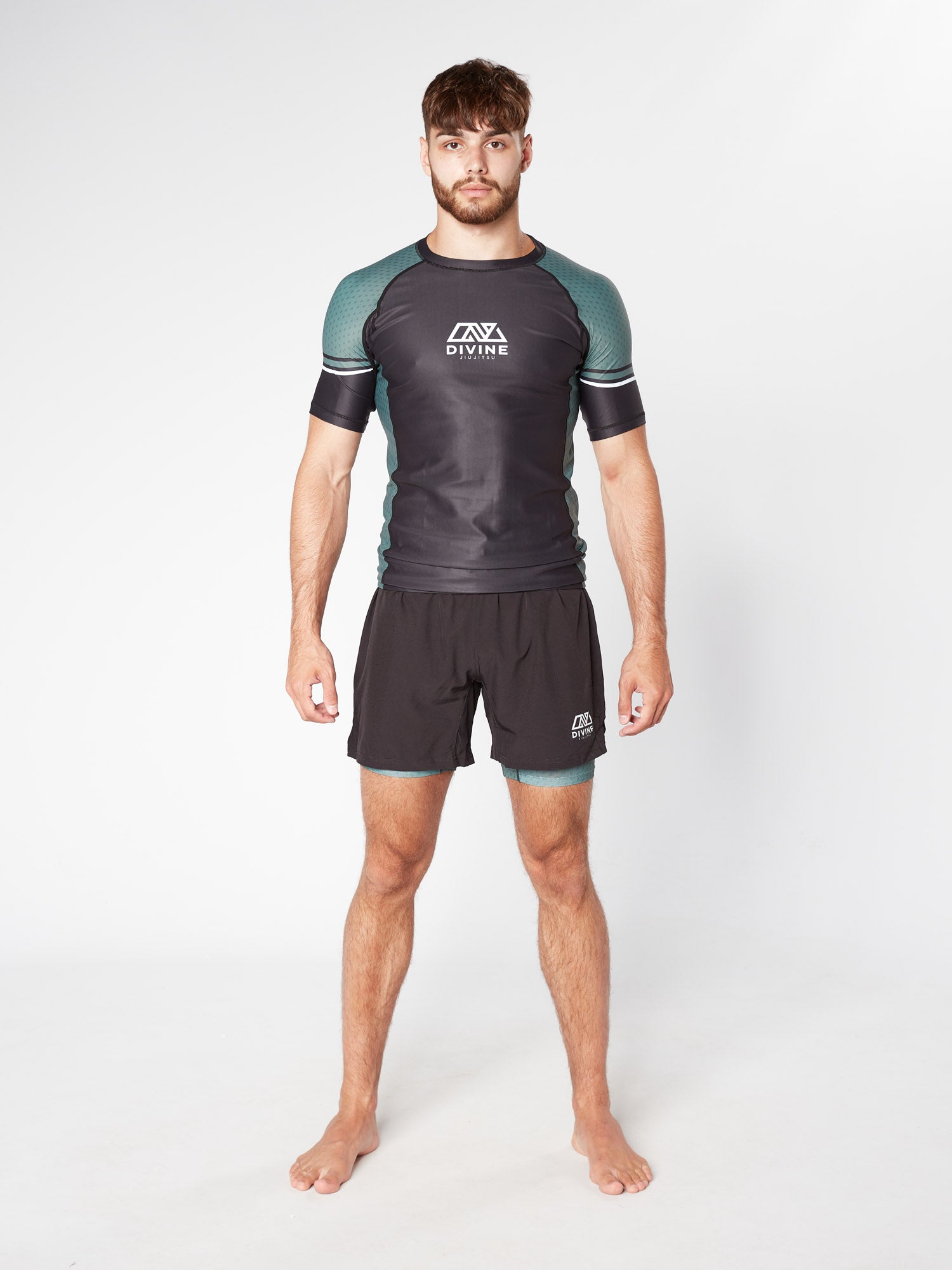 A man stands barefoot against a plain white background, wearing a fitted athletic outfit featuring the LEGION KHAKI BJJ RASHGUARD from Divine JiuJitsu in black and green and matching black shorts. Made from moisture-wicking fabric, his attire ensures optimum performance. His arms are relaxed at his sides, and he has a neutral expression.