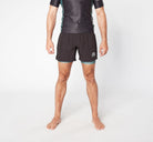 A person stands barefoot against a plain background, donning a black and dark teal short-sleeve compression shirt made from sustainable materials and Divine JiuJitsu's LEGION KHAKI BJJ SHORTS with turquoise mesh lining. Their hands are relaxed by their sides.