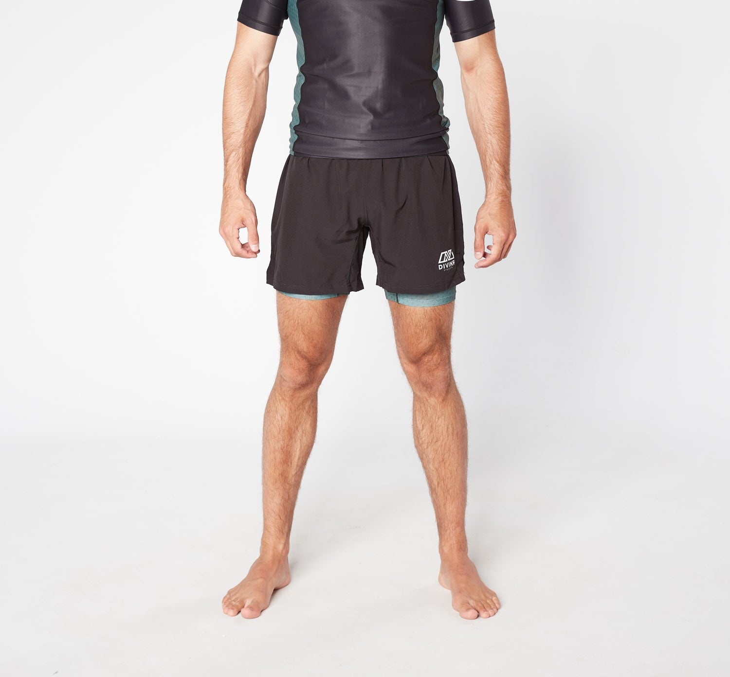 A person stands barefoot against a plain background, donning a black and dark teal short-sleeve compression shirt made from sustainable materials and Divine JiuJitsu's LEGION KHAKI BJJ SHORTS with turquoise mesh lining. Their hands are relaxed by their sides.