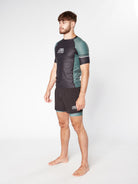 A bearded man with short hair stands barefoot in a studio against a plain white background, wearing the black and green LEGION KHAKI RASHGUARD by Divine JiuJitsu and matching shorts made of moisture-wicking fabric. He faces slightly to the side with a neutral expression.