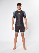 A man stands barefoot against a plain white background. He is wearing a tight Legion Grey bjj Rashguard by Divine JiuJitsu and matching black athletic shorts. His feet are slightly apart, and his arms hang naturally by his sides as he looks straight ahead.