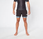 A person standing against a plain white background is wearing a black short-sleeve athletic shirt and Divine JiuJitsu Legion Grey bjj Shorts made from sustainable materials, with blue compression shorts underneath. They are barefoot, and their arms are relaxed at their sides.