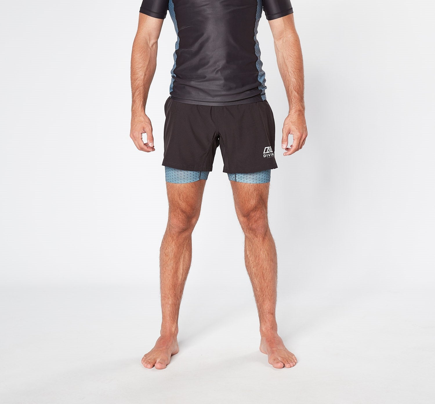 A person standing against a plain white background is wearing a black short-sleeve athletic shirt and Divine JiuJitsu Legion Grey bjj Shorts made from sustainable materials, with blue compression shorts underneath. They are barefoot, and their arms are relaxed at their sides.