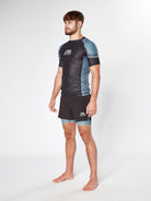 A barefoot man with a beard stands confidently, wearing a LEGION GREY RASHGUARD by Divine JiuJitsu, along with matching shorts. Both the shirt and shorts feature a hexagonal pattern and the logo "CV Fight Co." The background is plain white.