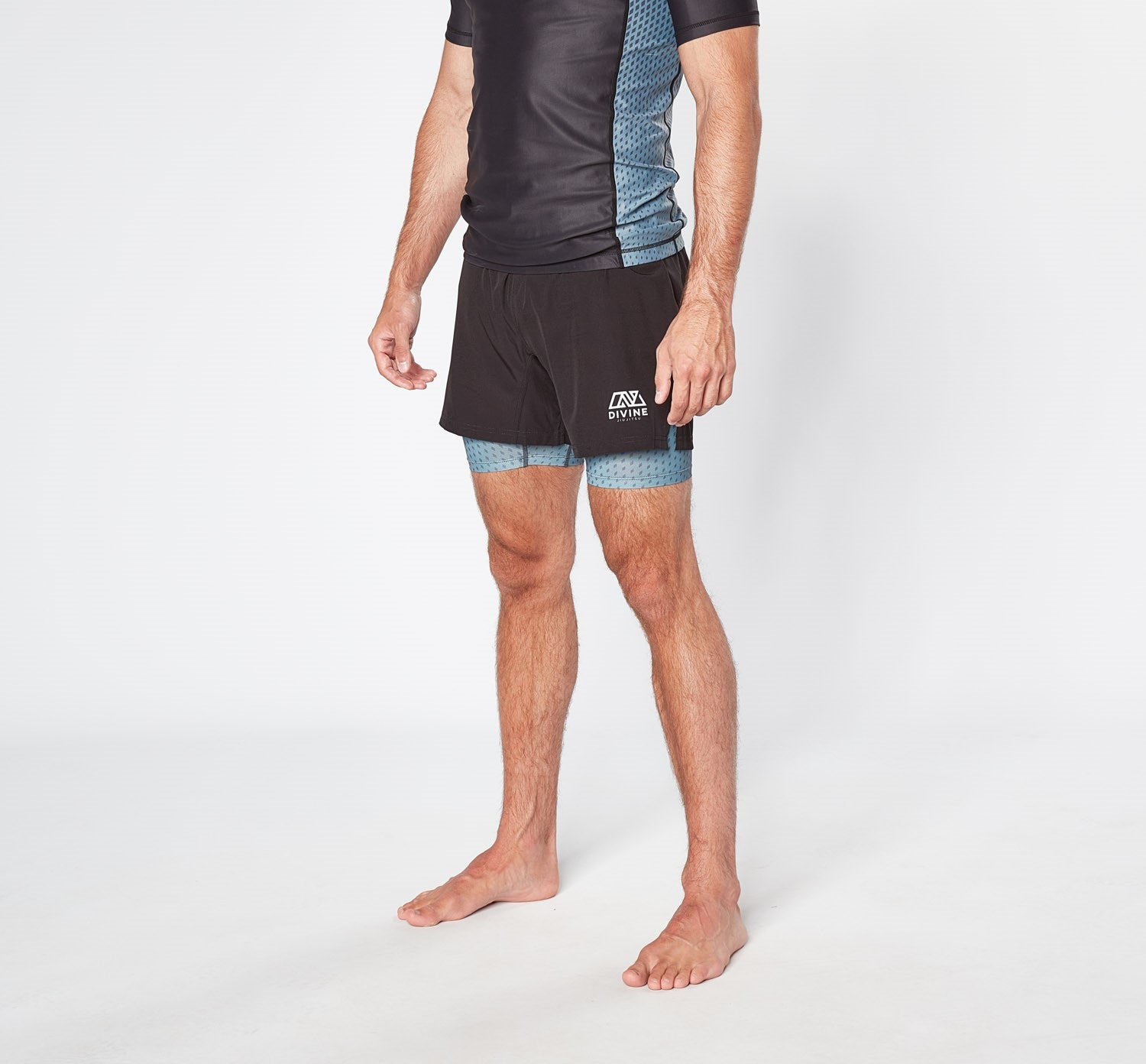 A person stands against a plain background wearing a black and blue short-sleeved athletic outfit. The LEGION GREY SHORTS by Divine JiuJitsu are made from quick-dry breathable fabric, featuring a blue pattern that extends slightly below the hem. The individual is barefoot, and the image is cropped above the shoulders.