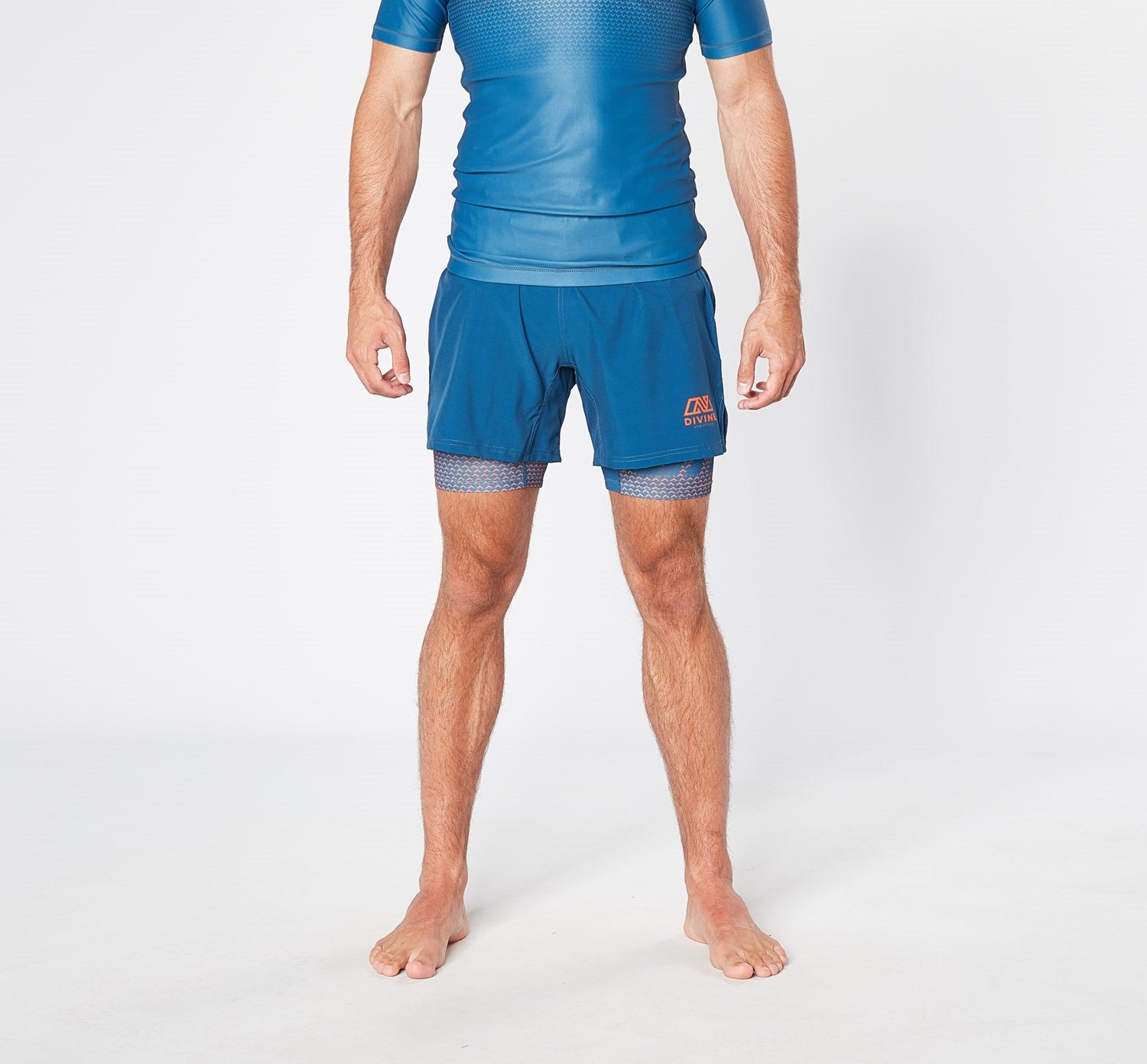 A man is standing in a studio setting, barefoot against a plain white background, wearing a fitted, quick-dry blue athletic shirt and the ILLENIUM BJJ SHORTS by Divine JiuJitsu in blue with inner leggings. The anti-wrinkle fabric of his attire ensures he looks sharp, and the eco-friendly material makes it a conscious choice.