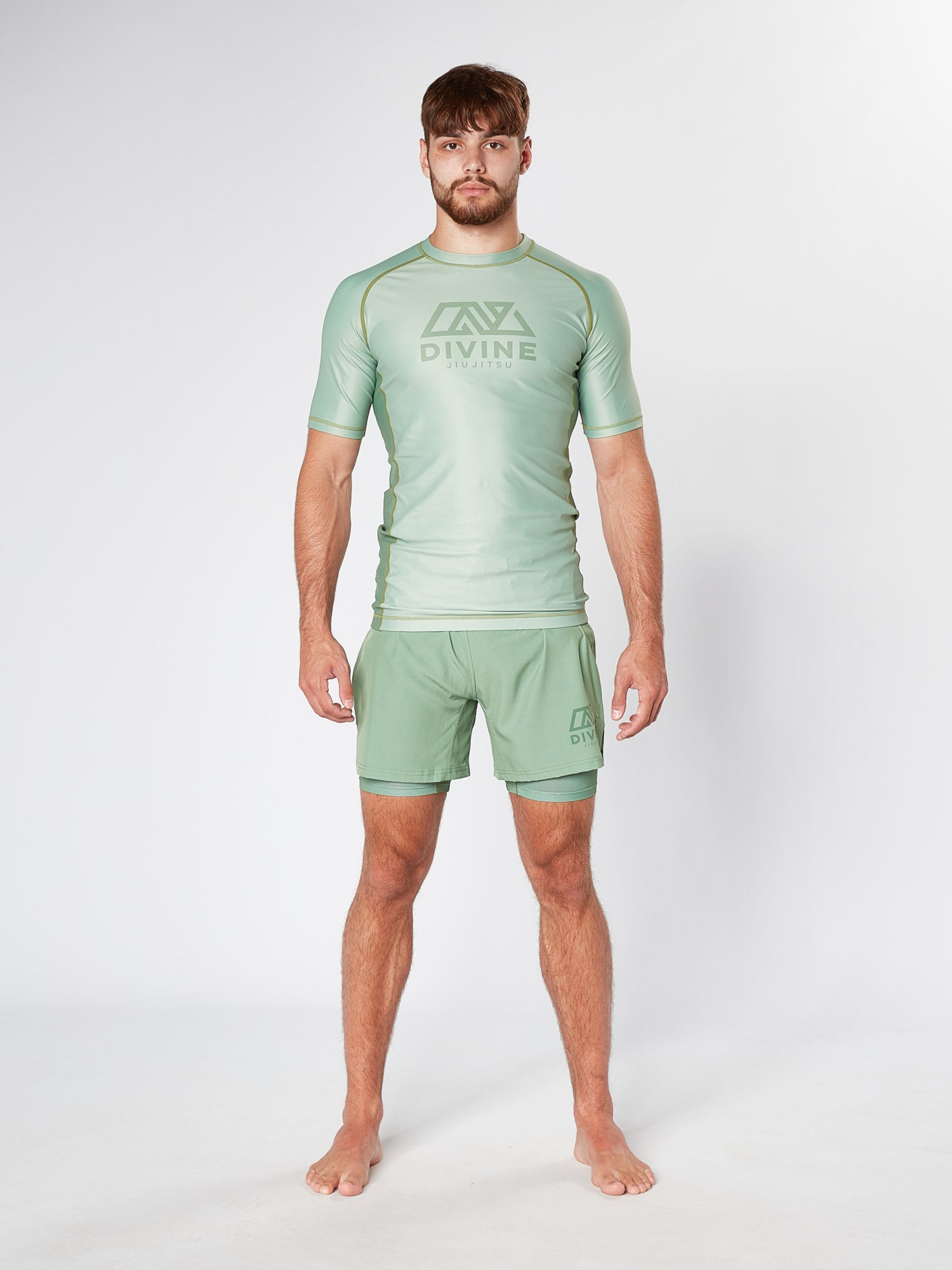 A man stands barefoot against a plain background, wearing a matching sage green KENJA BJJ RASHGUARD athletic set from Divine JiuJitsu that includes a short-sleeve shirt and shorts. Both pieces feature the "DIVINE" logo along with a stylized graphic. His hair is short, he sports a beard, and the outfit resembles jiu jitsu rashguard attire perfect for martial arts training.