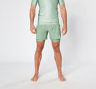 A person with short hair stands barefoot on a white surface, wearing a light green, short-sleeved athletic top and matching KENJA BJJ SHORTS from Divine JiuJitsu. The photo captures the individual from the neck down, highlighting their sage green sports attire.