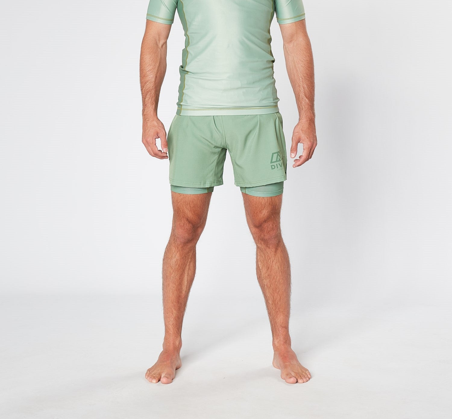 A person with short hair stands barefoot on a white surface, wearing a light green, short-sleeved athletic top and matching KENJA BJJ SHORTS from Divine JiuJitsu. The photo captures the individual from the neck down, highlighting their sage green sports attire.