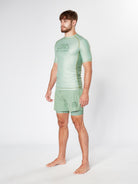 A man stands barefoot against a plain white background, wearing a sage green KENJA BJJ RASHGUARD and matching shorts. The athletic wear, crafted for martial arts aficionados, features the logo and name "Divine JiuJitsu" on both the shirt and shorts.