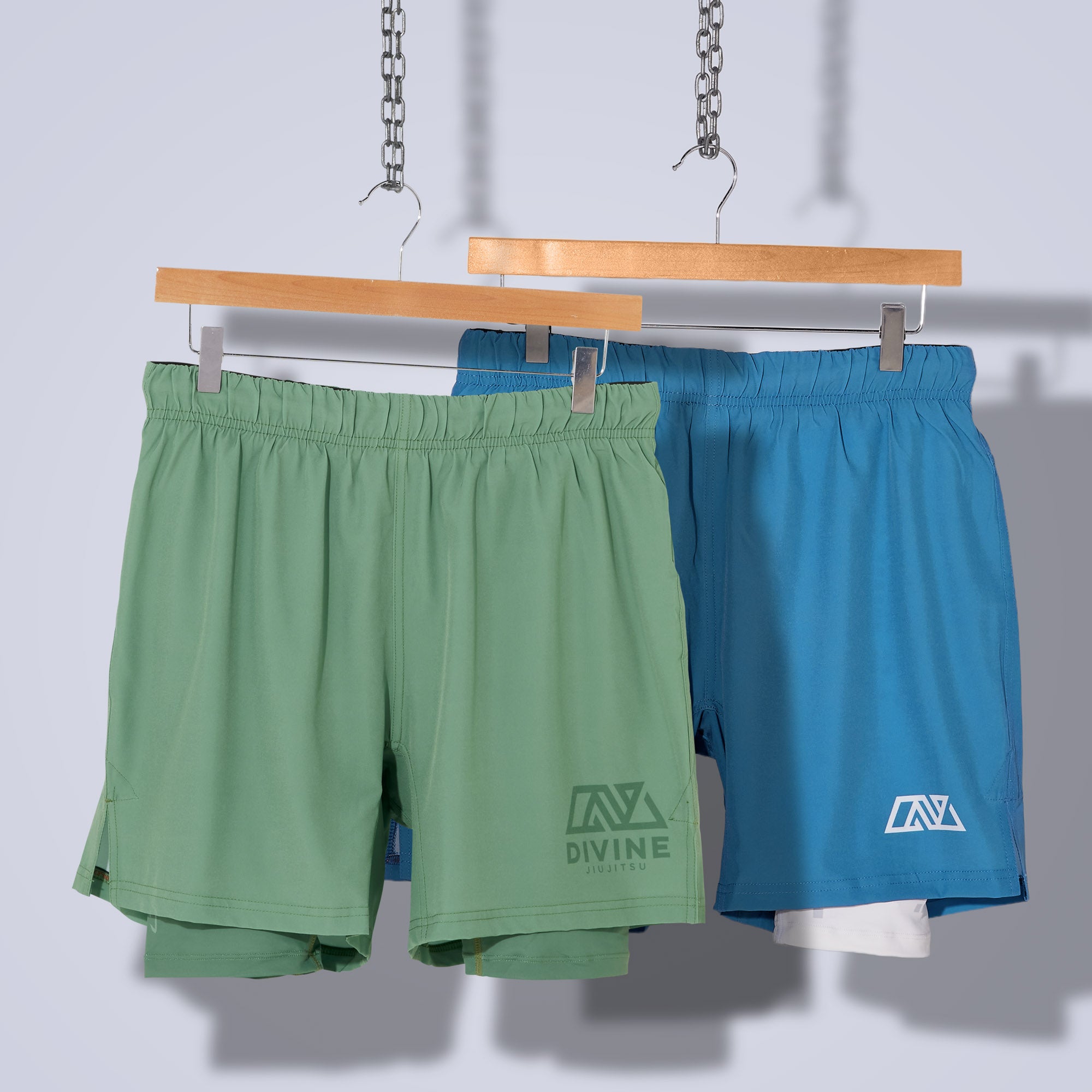 Two pairs of athletic shorts hanging on wooden hangers suspended by metal chains. One pair is green with the "Divine" brand logo, and the other is blue with a white inner layer showing at the bottom and a similar logo. Both have front pockets.