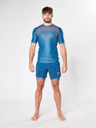 A barefoot man stands against a plain backdrop, wearing the ILLENIUM BJJ RASHGUARD from Divine JiuJitsu in blue and orange, with "DIVINE" text, along with matching shorts. He has a serious expression and a well-built physique.