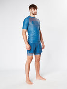 A man with short hair and a beard stands barefoot on a neutral background, wearing the ILLENIUM RASHGUARD athletic set from Divine JiuJitsu, consisting of a blue short-sleeve top and blue shorts. The jiu-jitsu enthusiast faces slightly to the side, his arms relaxed by his sides, embodying both performance and style.