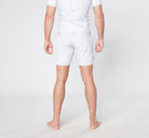 A person stands barefoot on a light-colored floor, seen from behind. They are wearing a white athletic outfit from Divine JiuJitsu, featuring BARE BLANCO BJJ SHORTS and a matching short-sleeve shirt made from quick dry breathable fabric. The background is plain and white.