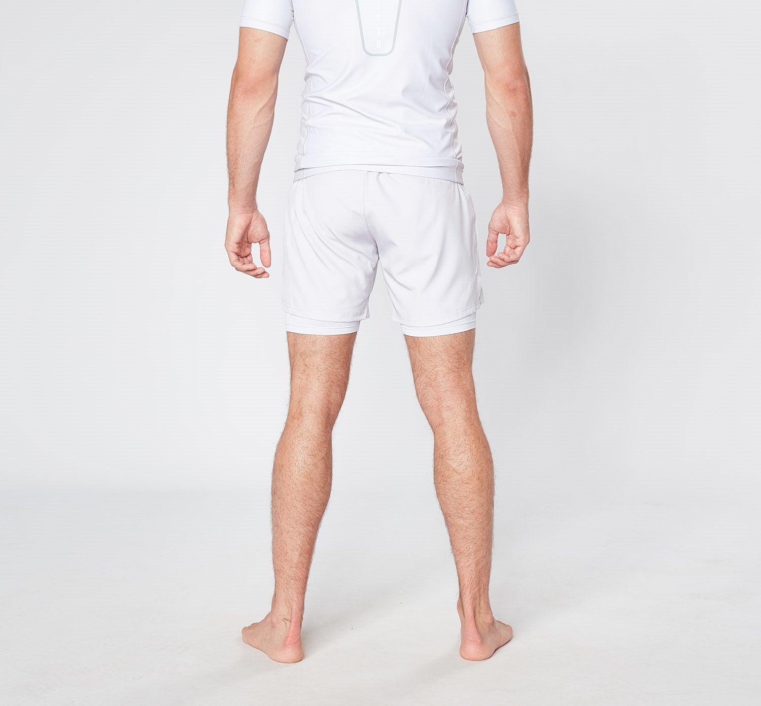 A person stands barefoot on a light-colored floor, seen from behind. They are wearing a white athletic outfit from Divine JiuJitsu, featuring BARE BLANCO BJJ SHORTS and a matching short-sleeve shirt made from quick dry breathable fabric. The background is plain and white.