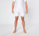 A person is standing barefoot against a plain background, wearing a white short-sleeved athletic shirt and the BARE BLANCO BJJ SHORTS from Divine JiuJitsu, made from sustainable materials. The person’s arms are relaxed by their sides, and their legs are slightly apart. Their face is not visible in the image.