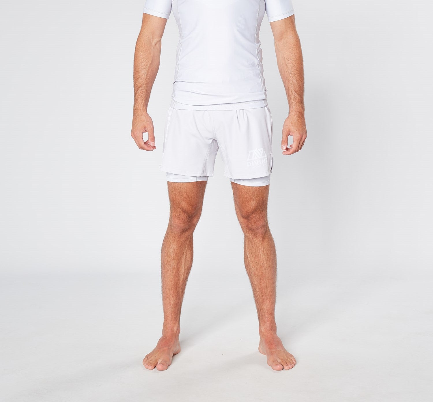 A person is standing barefoot against a plain background, wearing a white short-sleeved athletic shirt and the BARE BLANCO BJJ SHORTS from Divine JiuJitsu, made from sustainable materials. The person’s arms are relaxed by their sides, and their legs are slightly apart. Their face is not visible in the image.