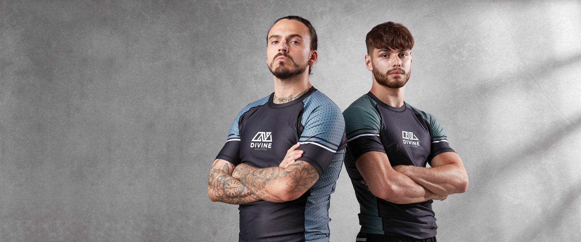 Two men standing with arms crossed, wearing athletic gear with the logo "AVLDIVINE" on their shirts. The man on the left has tattoos on both arms and a bun hairstyle, while the man on the right has short hair and a beard. The background is a plain, textured gray wall.